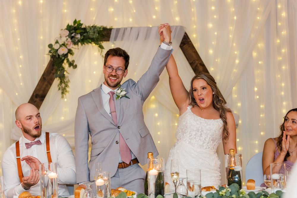 Wedding party celebrates during reception with twinkle lights and decor in background.