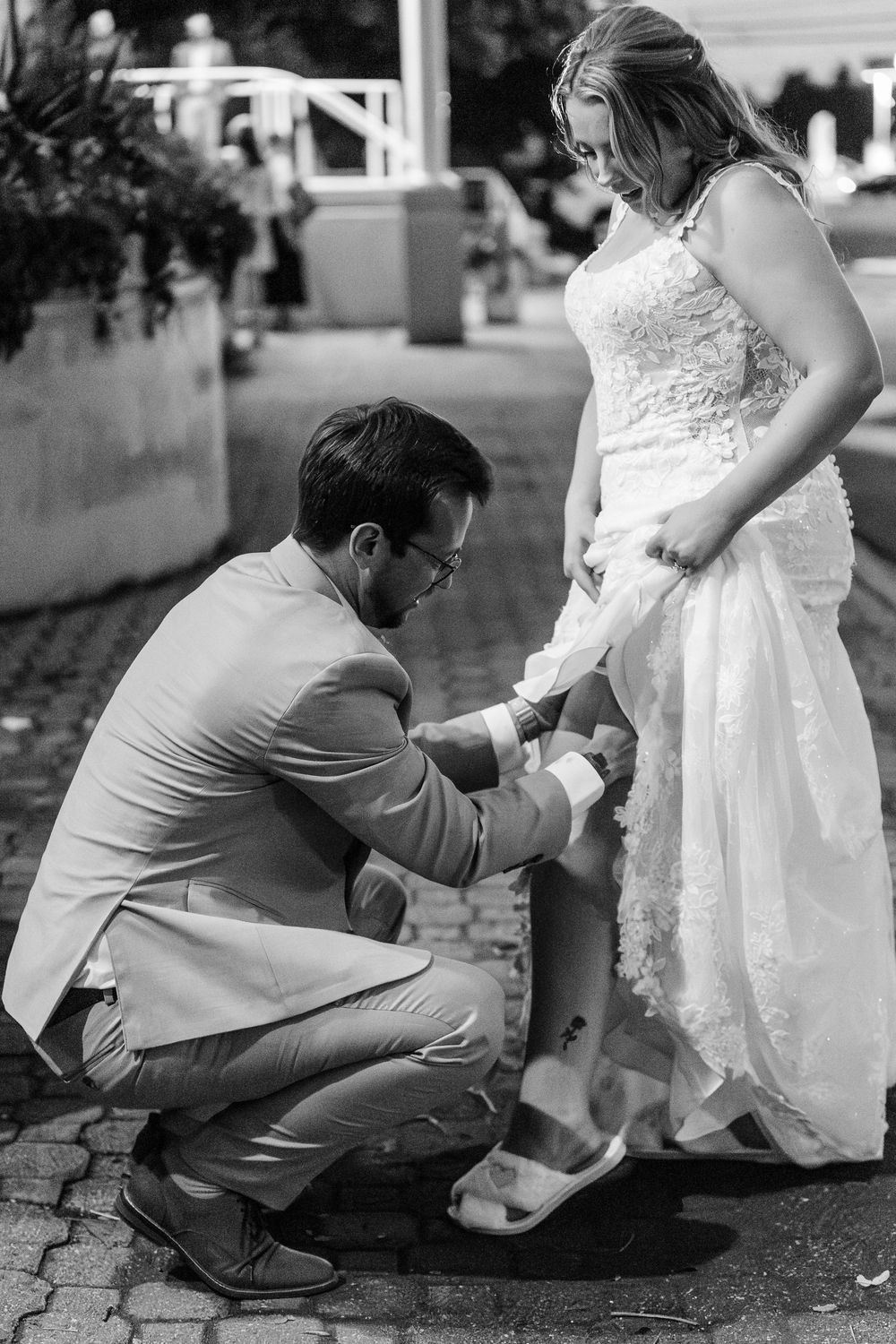 A romantic monochromatic sequence shows a groom helping his bride adjust her shoe on the stone pavement.