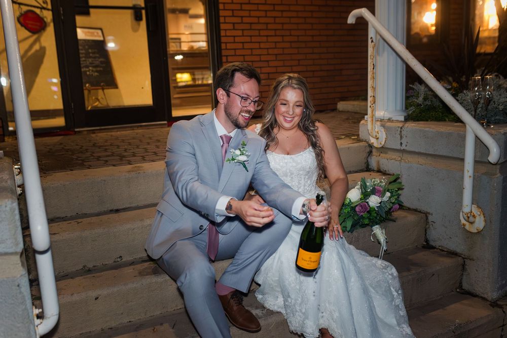 Wedding party members in gray suits gather at a venue entrance while holding champagne bottles and celebrating.