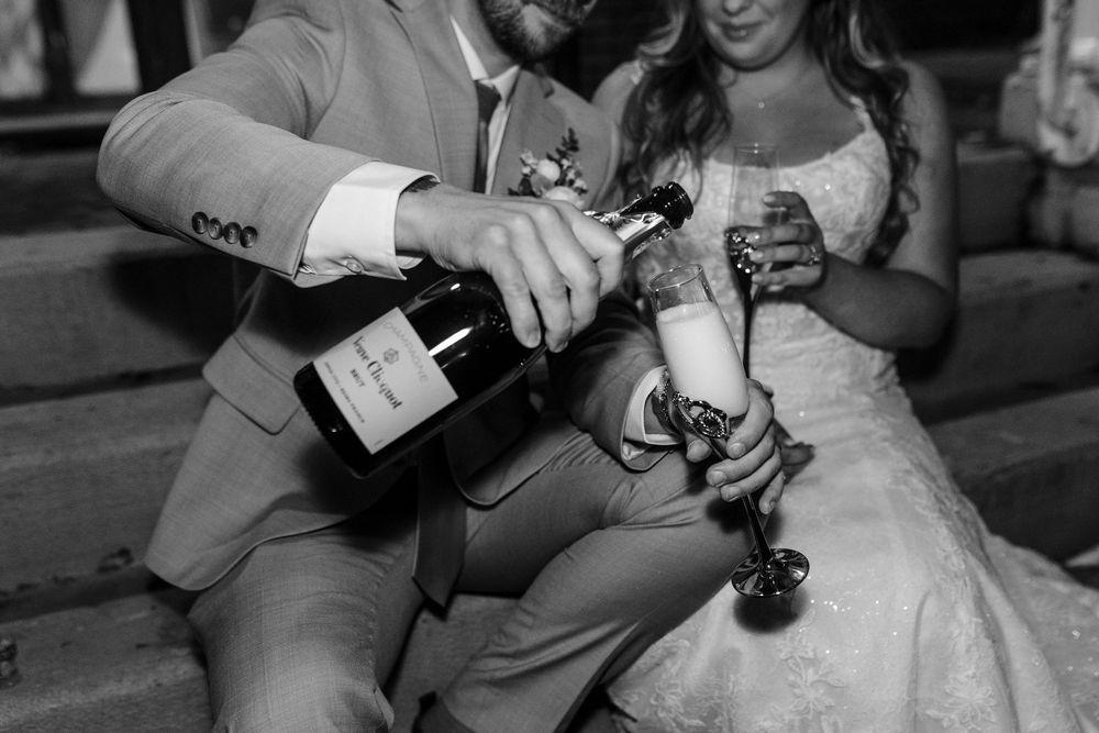 Black and white photo of a couple pouring champagne into glasses during a celebratory moment at their wedding.