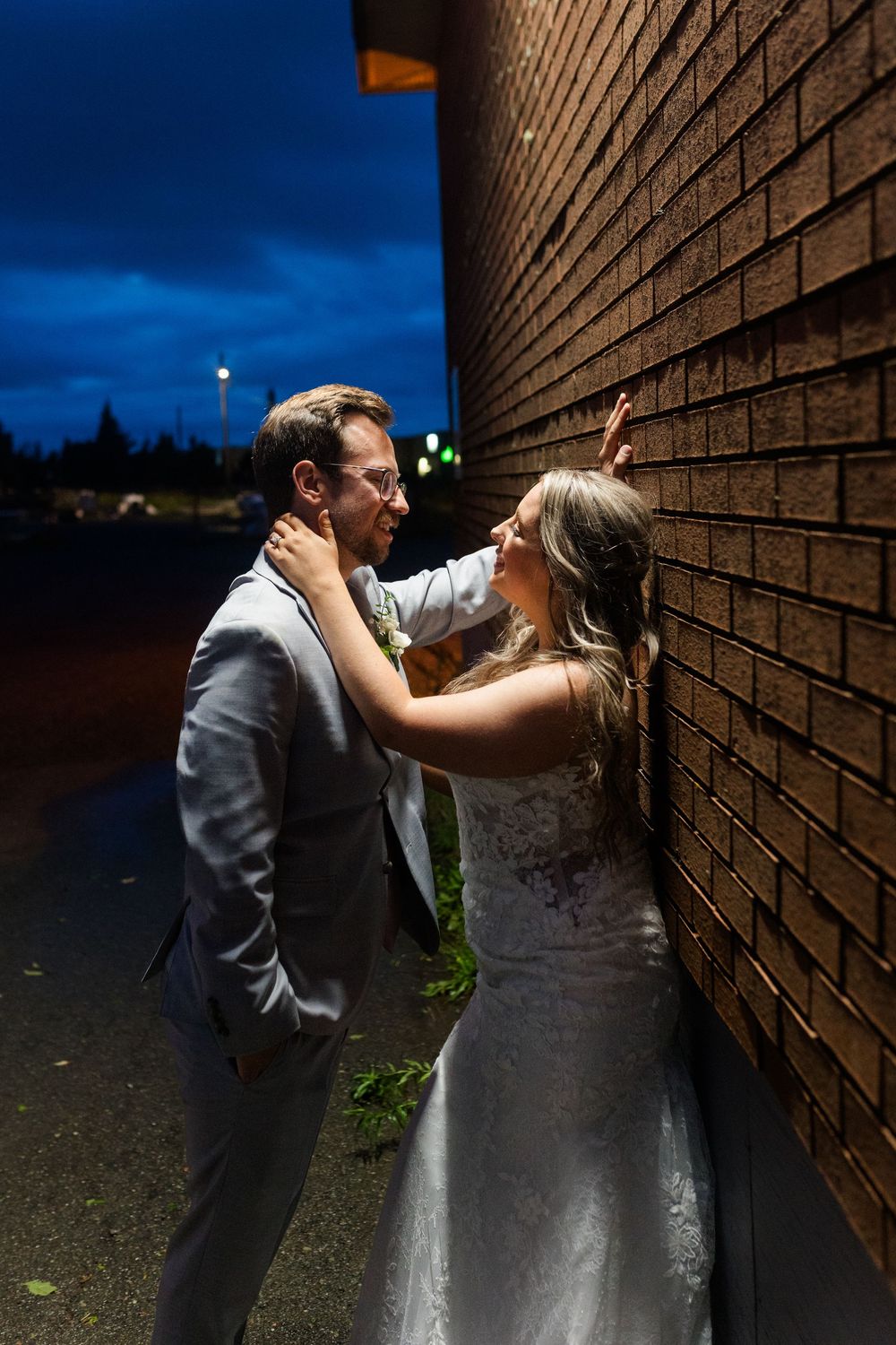 A romantic newlywed couple shares an intimate moment against a brick wall in the evening light.