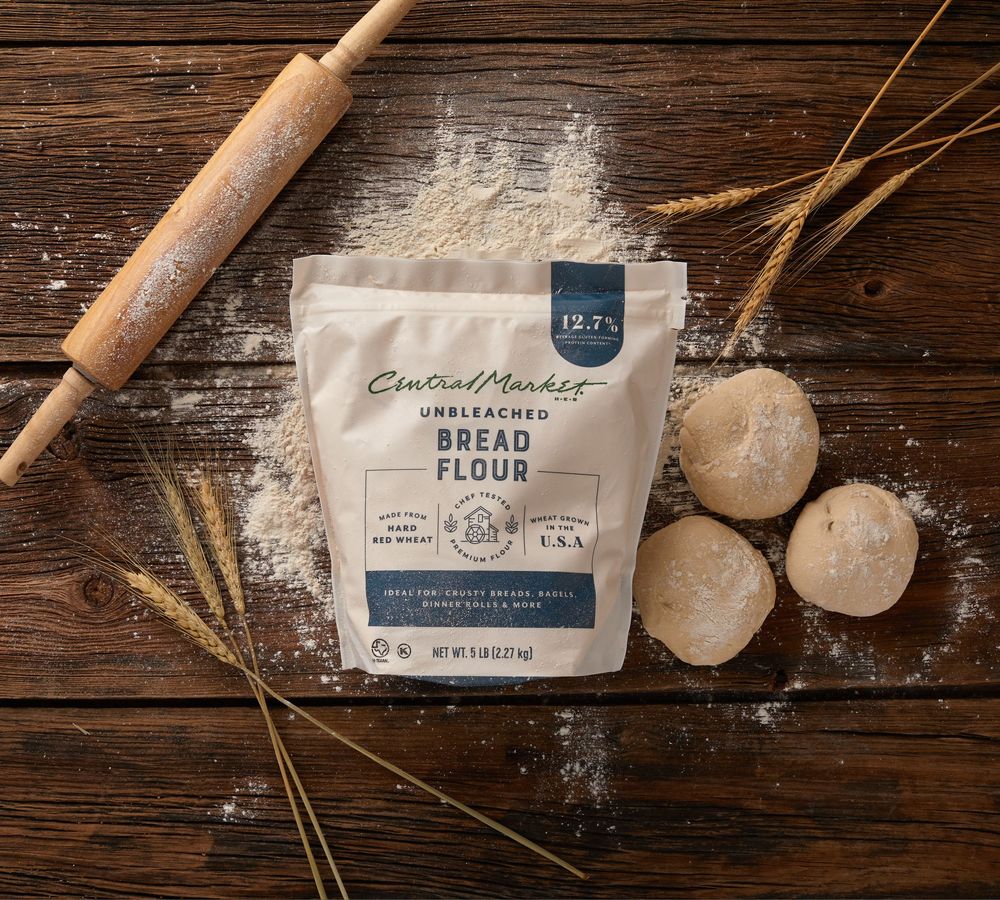 Rustic baking ingredients including flour, dough, and wheat stalks arranged on weathered wooden surface with natural lighting.
