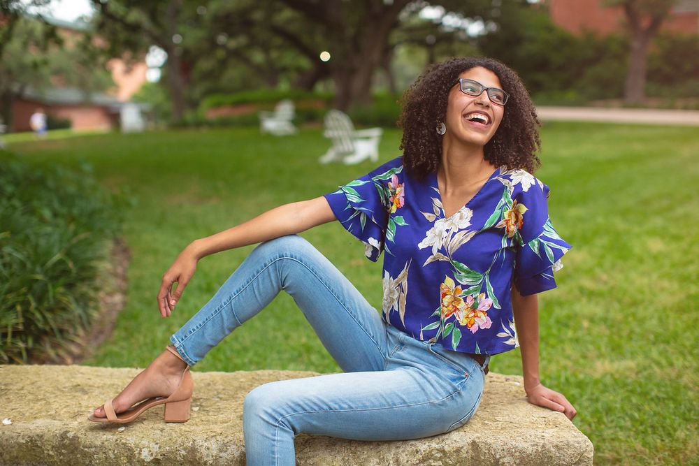 Woman in colorful top and jeans laughs joyfully while sitting on grass.
