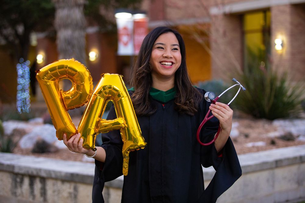 Graduate holding gold letter balloons and diploma during graduation celebration at dusk.