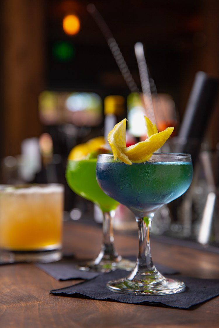 Colorful cocktails with fruit garnishes served in martini glasses on dark coasters at an upscale bar.