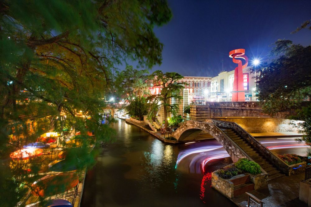 Scenic night view of city riverfront with illuminated bridges, buildings and trees reflecting in calm water.