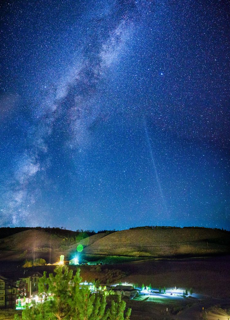 Stunning night sky with Milky Way galaxy visible over illuminated buildings in rural landscape.