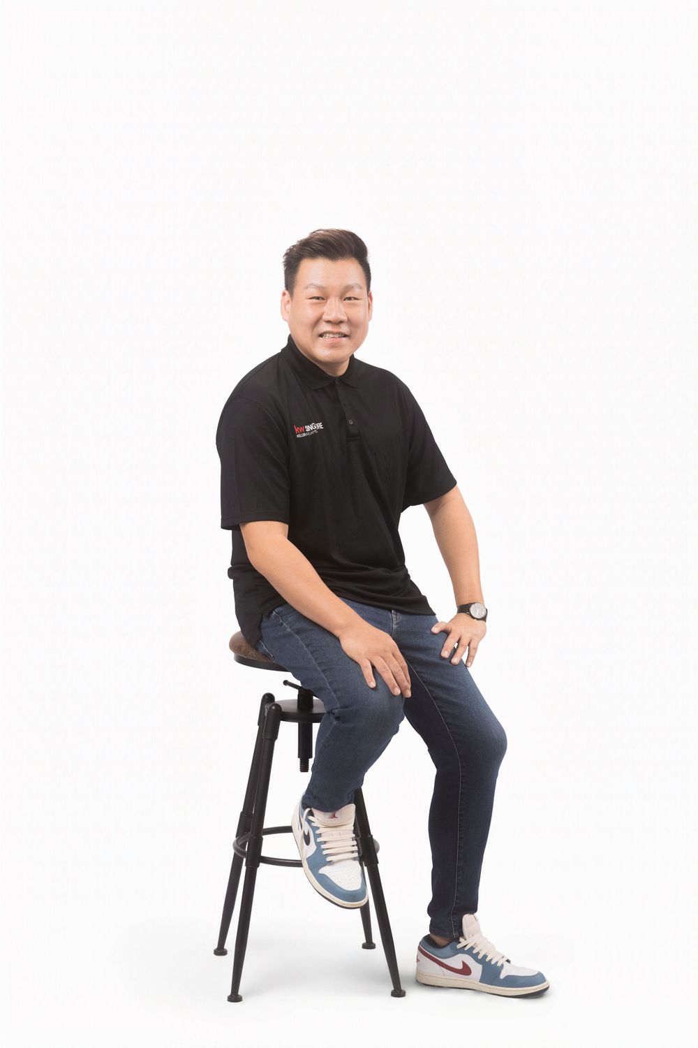 Person in black polo shirt and jeans sitting on bar stool against white background.