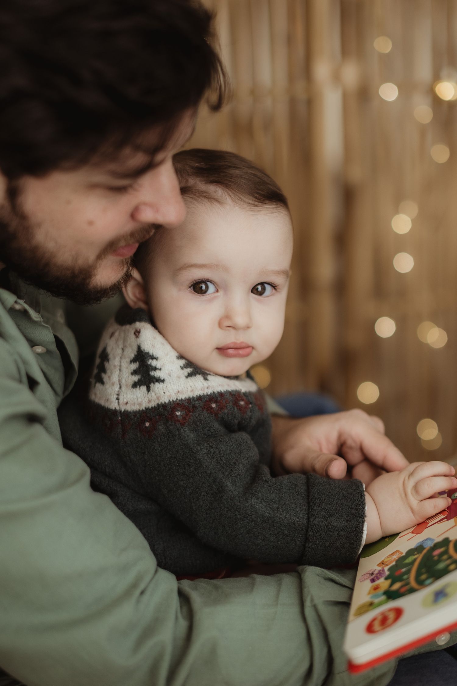 Parent and child share intimate reading moment near Christmas lights backdrop.