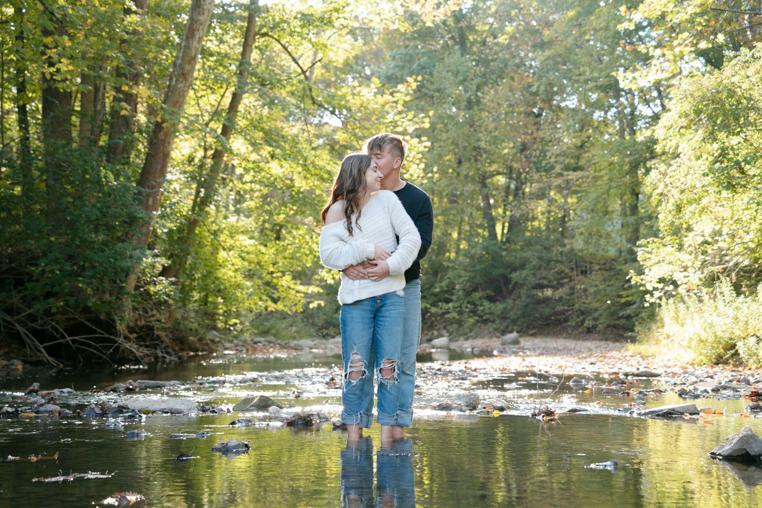 Couple embracing and kissing while standing in shallow creek surrounded by lush green forest trees.