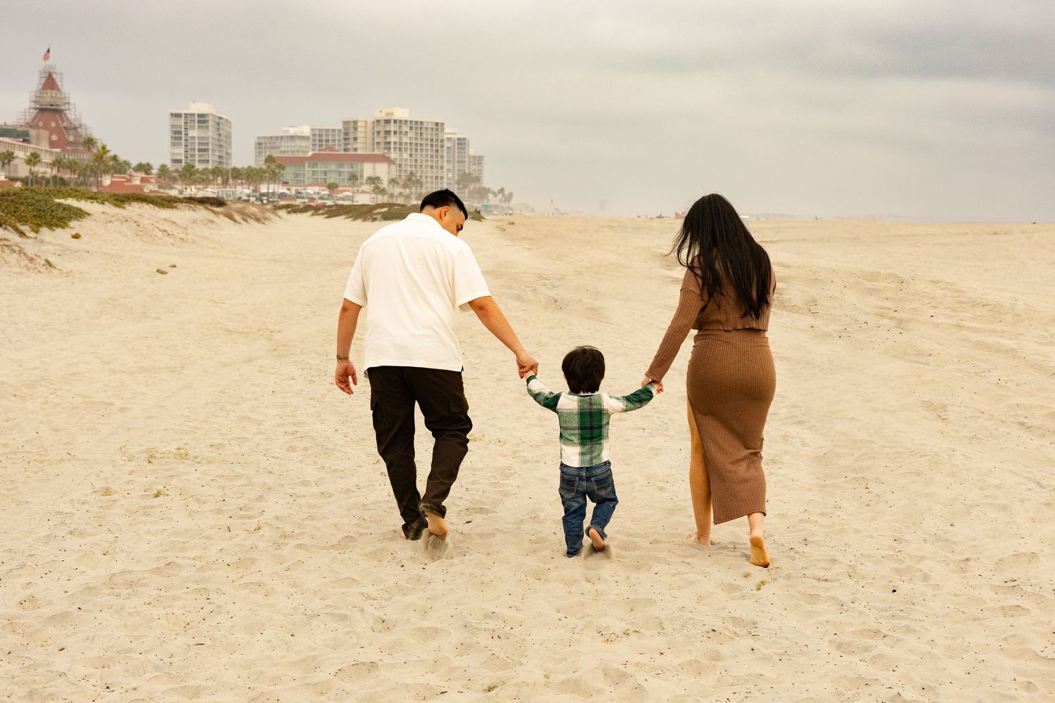 Parents and child holding hands while walking on the sand at Coronado Beach. San Diego family photographer