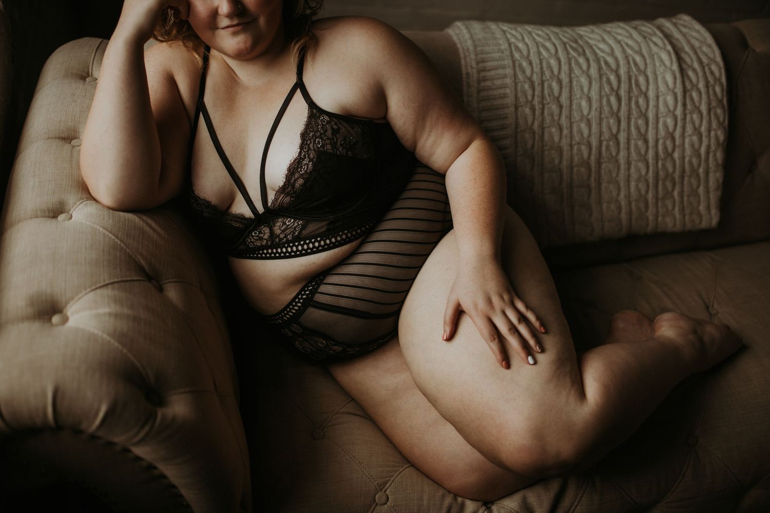 A seated figure in moody natural lighting shows off their shoulder tattoos while sitting on a bed.