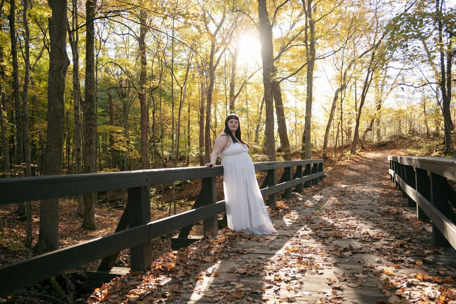 Woman in white flowing gown standing on wooden bridge surrounded by sunlit autumn forest foliage.