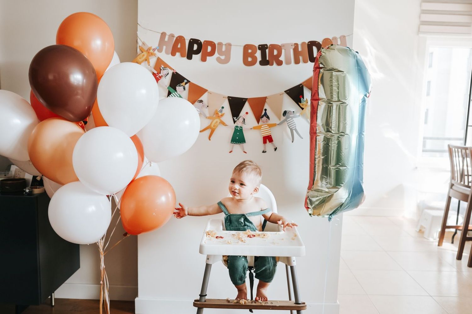 First birthday celebration setup with orange and white balloons, banner and high chair against white wall.