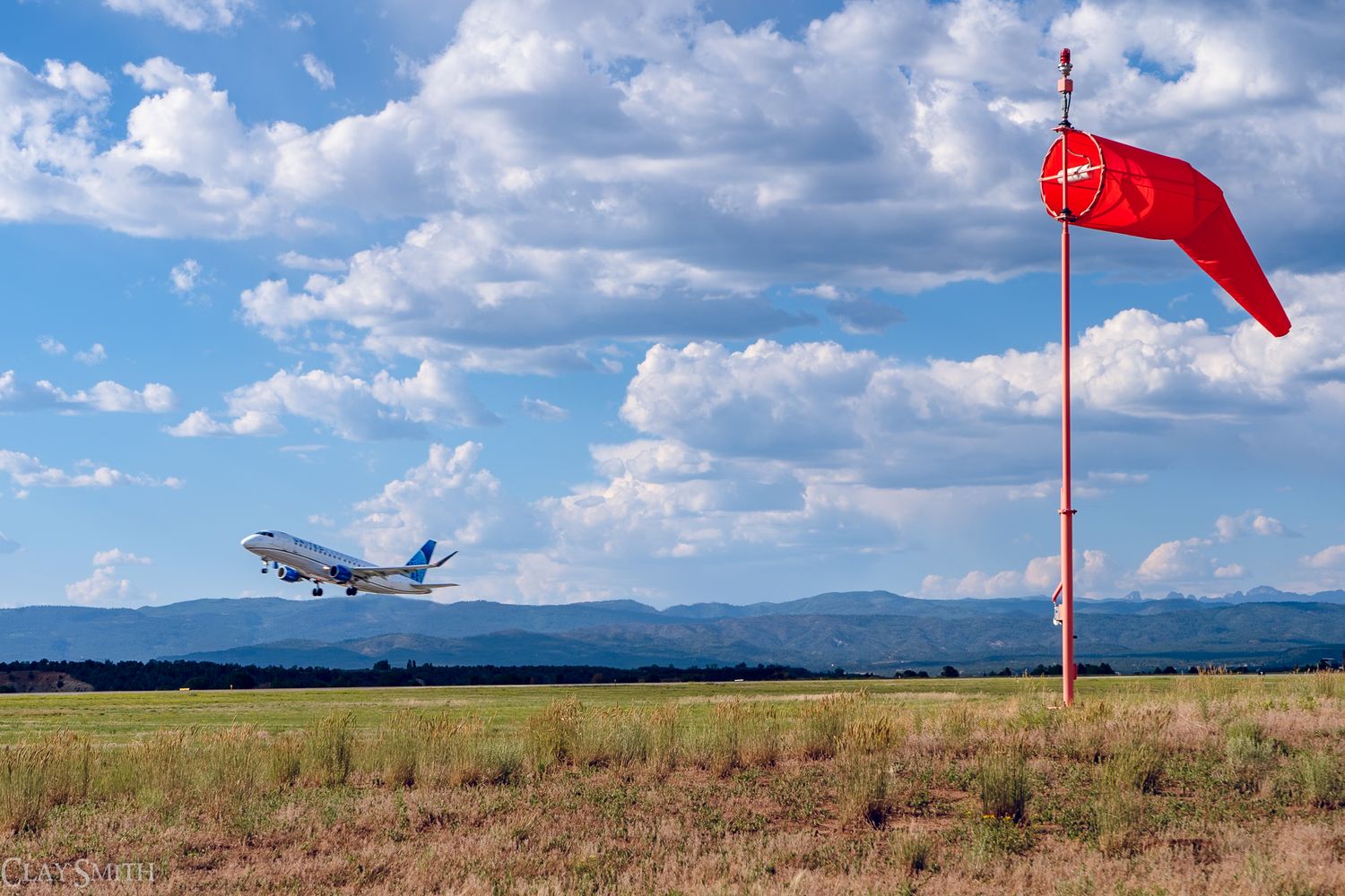 A commercial airplane takes off near a red wind sock with mountains and puffy white clouds in the background.