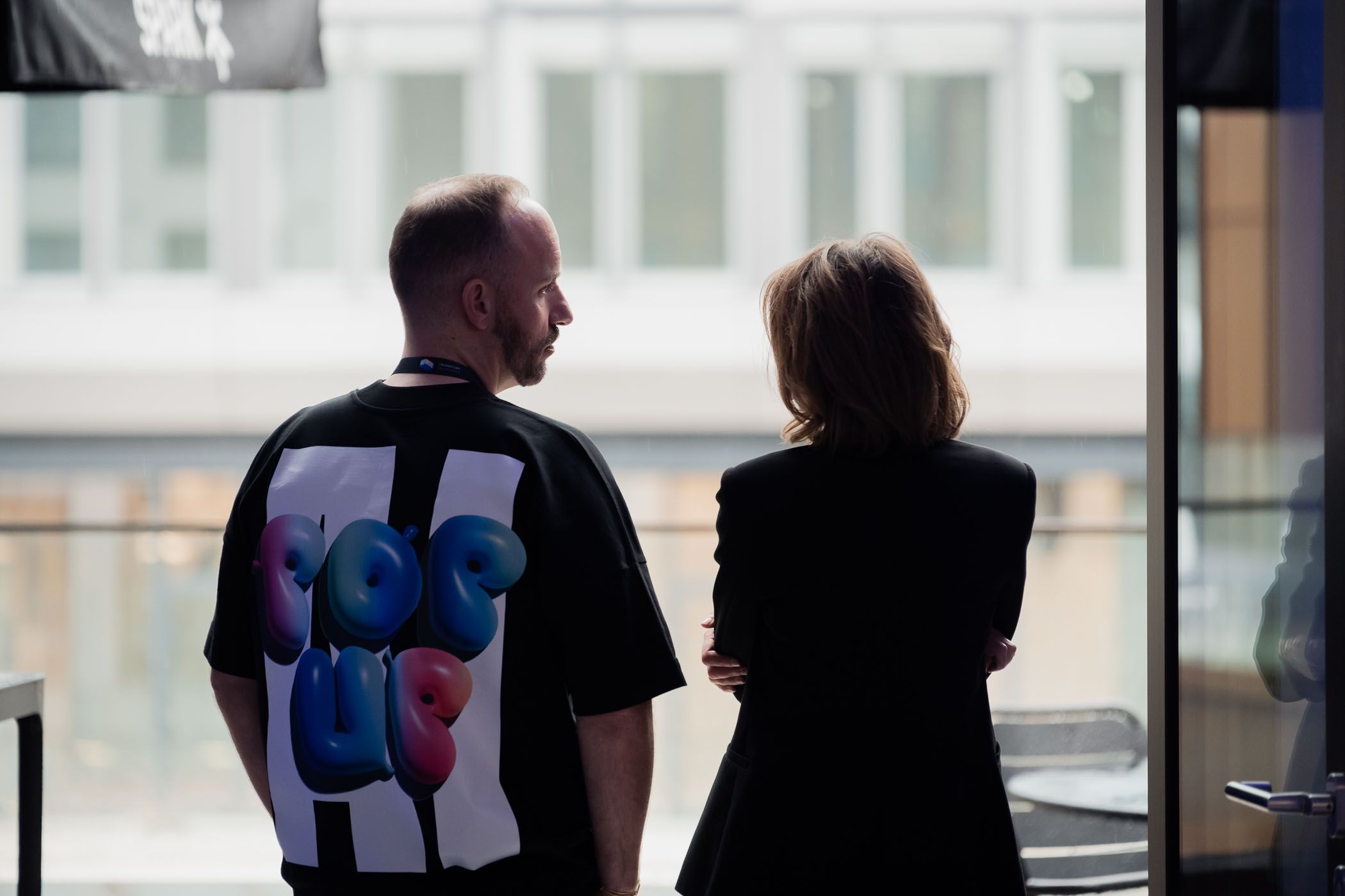 Two people in silhouette having a conversation near large windows.