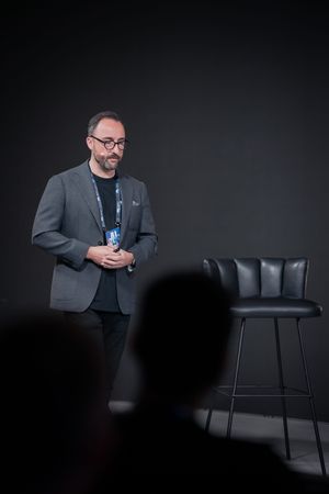 Speaker in gray blazer presenting against dark background with bar stool.