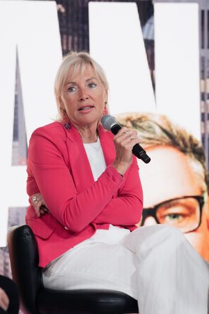 Professional speaker in pink blazer presenting at conference with cityscape backdrop.