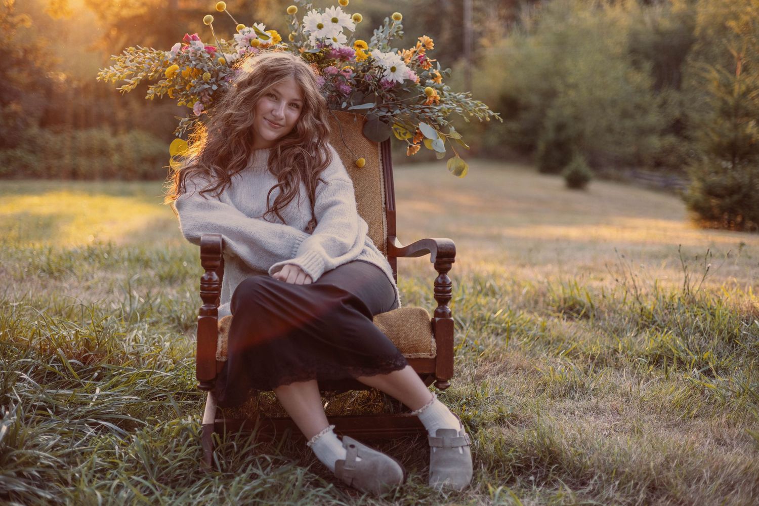 Portrait of woman in white sweater sitting in vintage chair with floral crown outdoors.