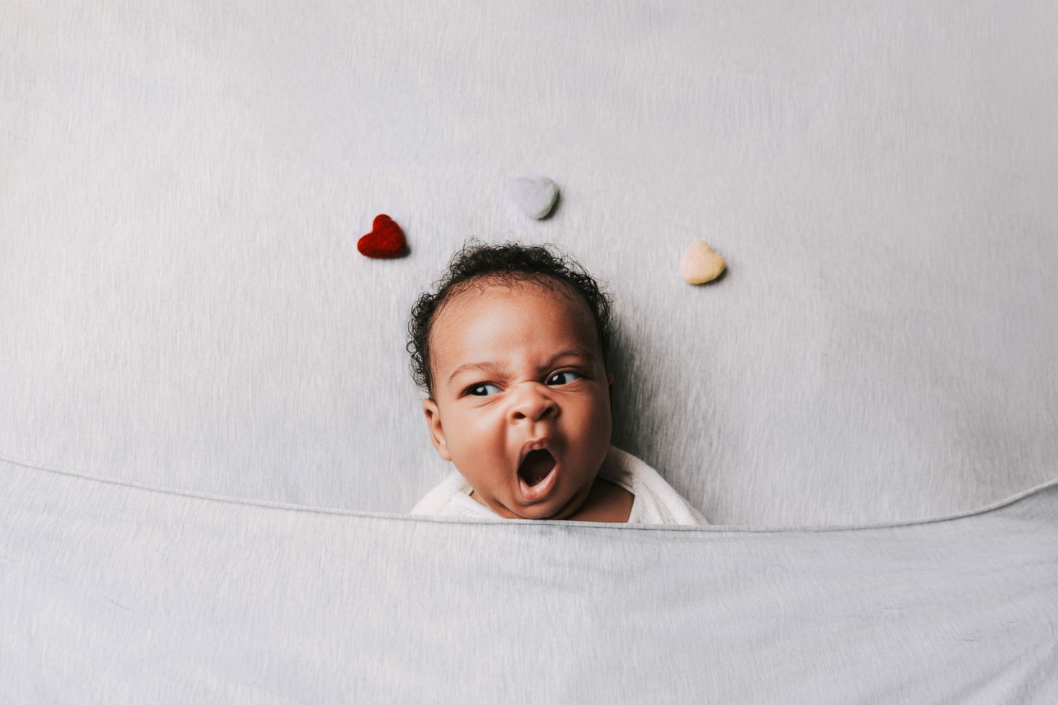 Adorable Black baby yawning while lying in a white blanket, surrounded by colorful heart decorations.