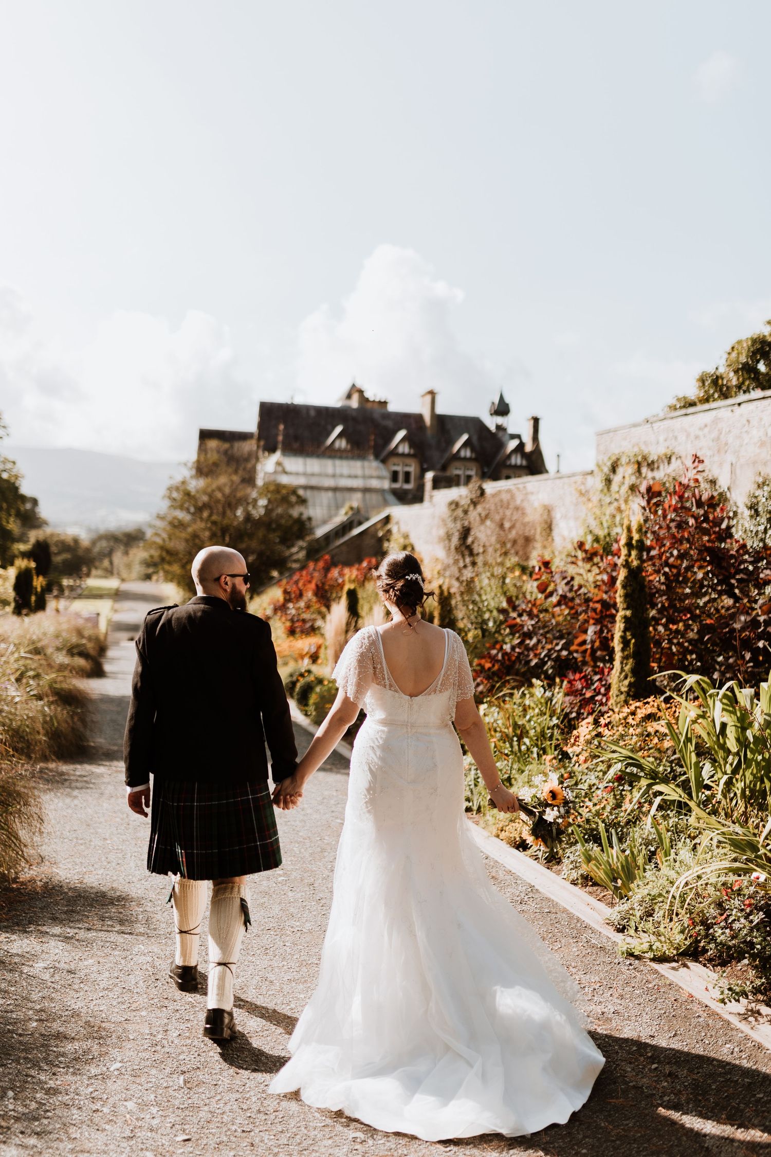 A couple walks along a garden path at a Scottish wedding, with the groom in traditional kilt and bride in flowing white gown.
