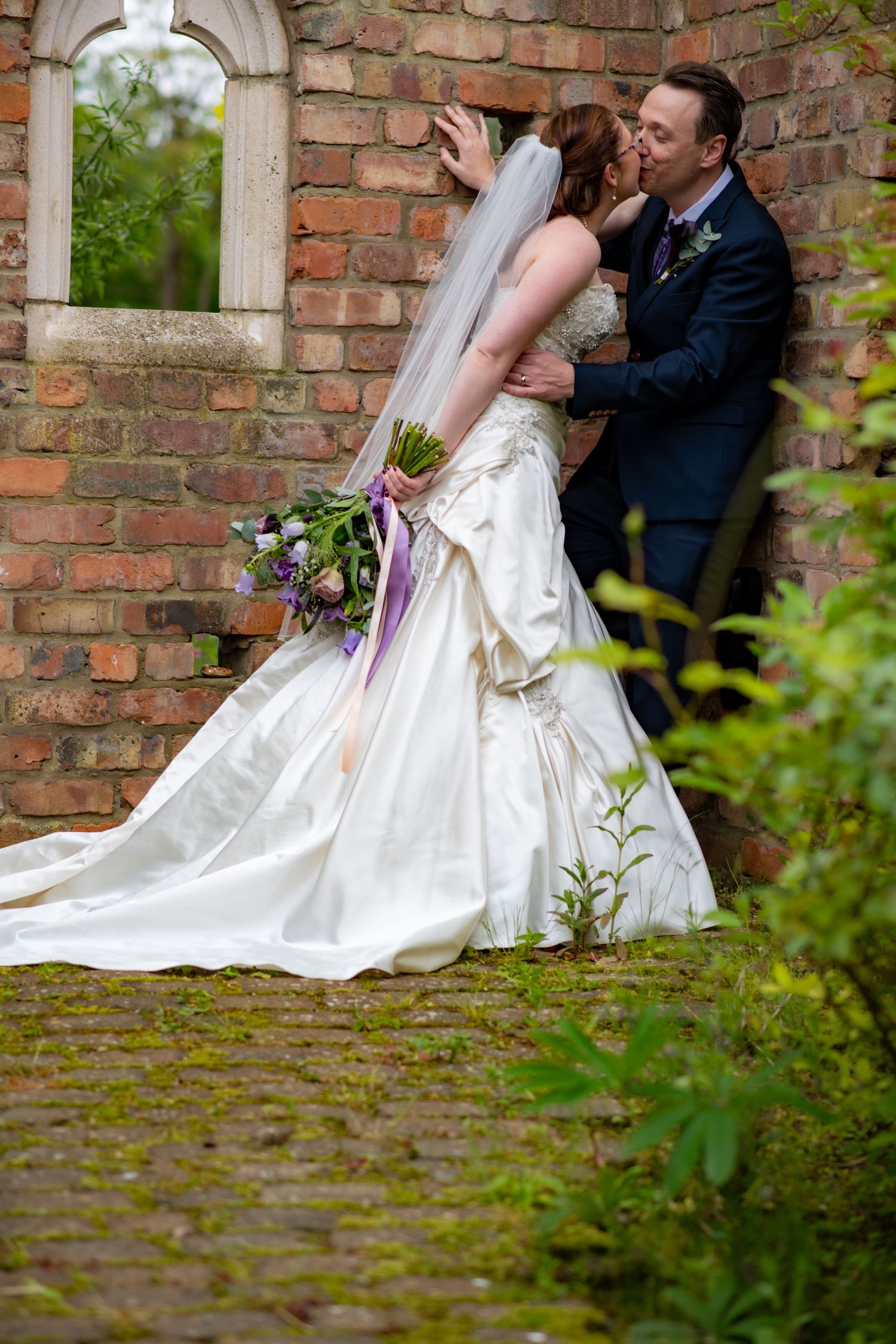 Wedding couple sharing romantic moment by brick archway with trailing flowers.
