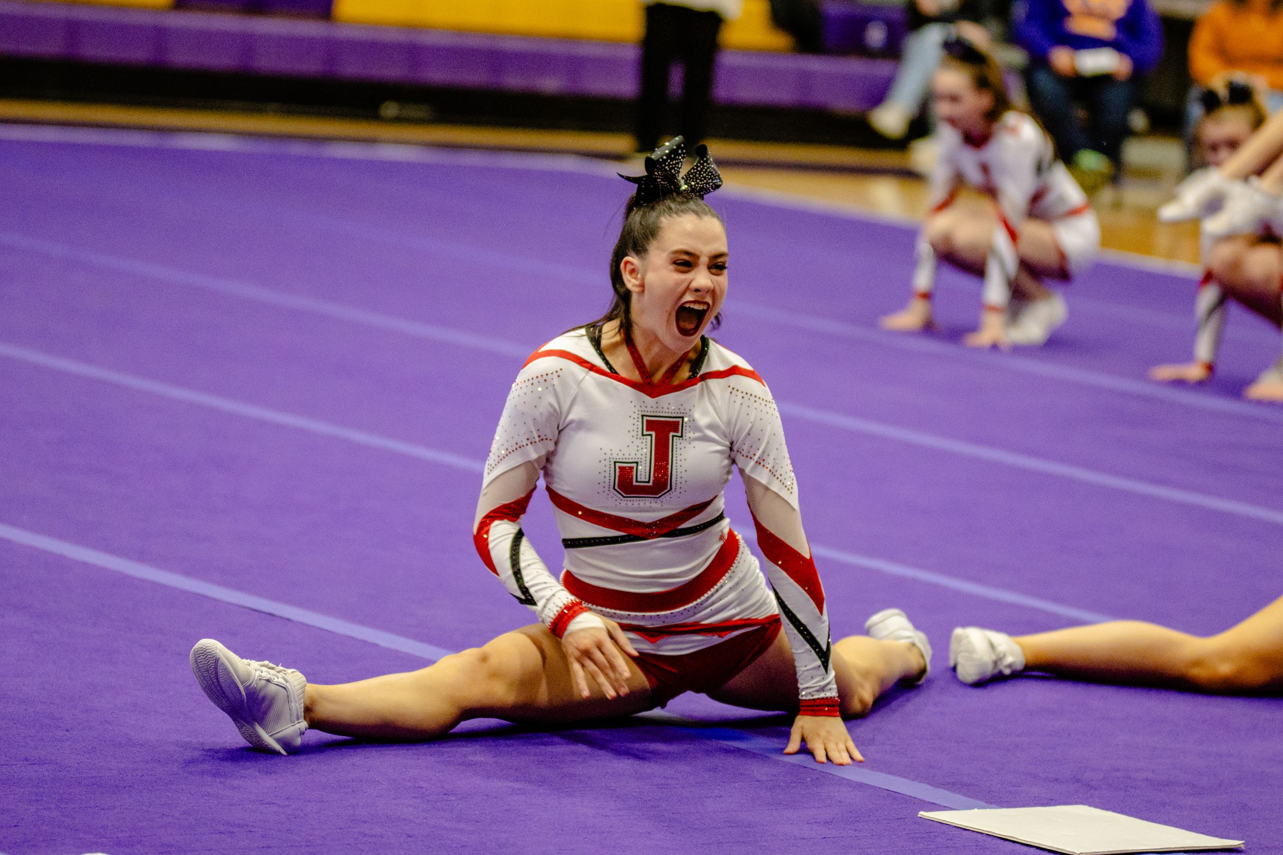 Cheerleader in uniform performs splits during competition on purple mat.