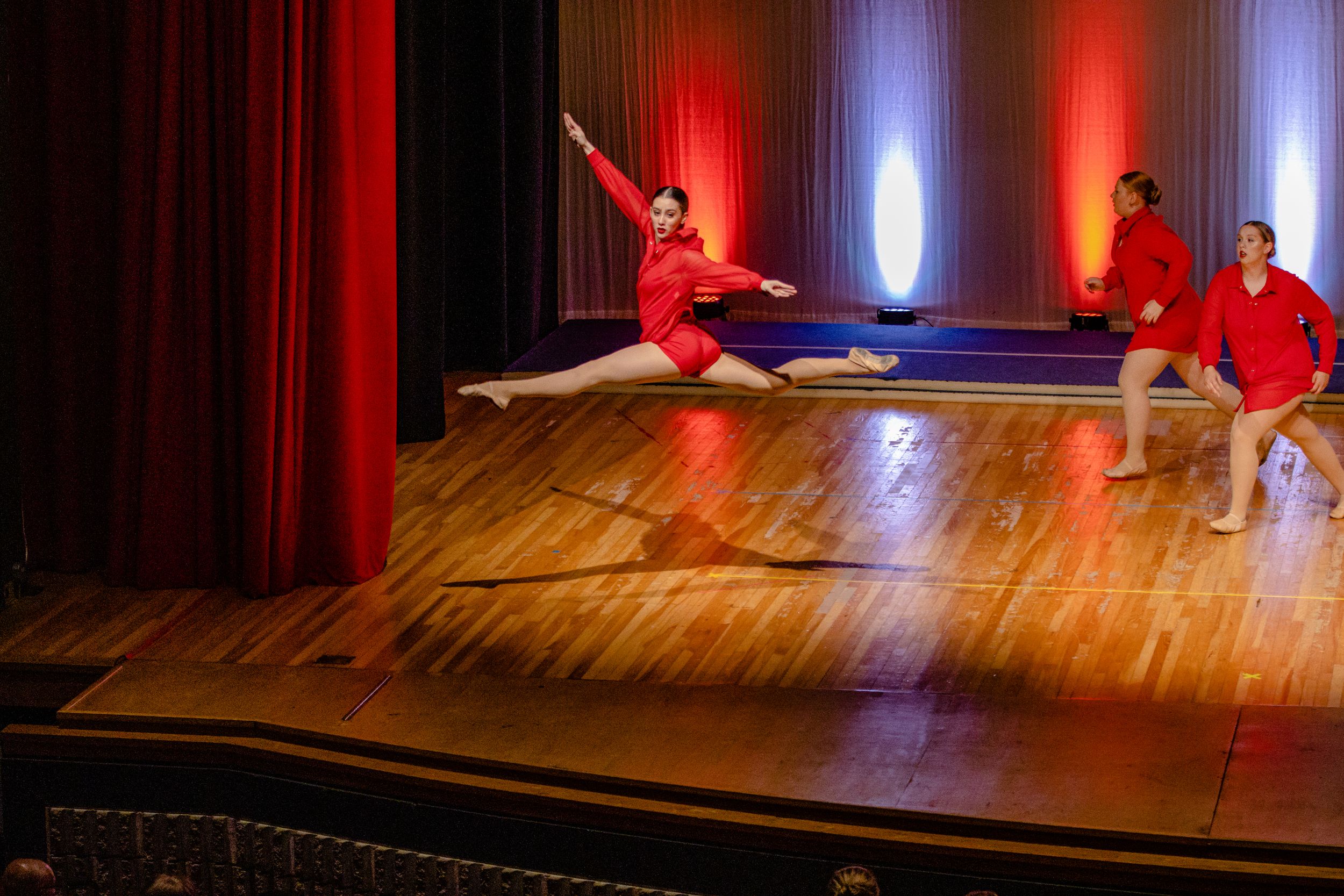 Dancers in red costumes perform a dynamic leap sequence on a wooden stage.