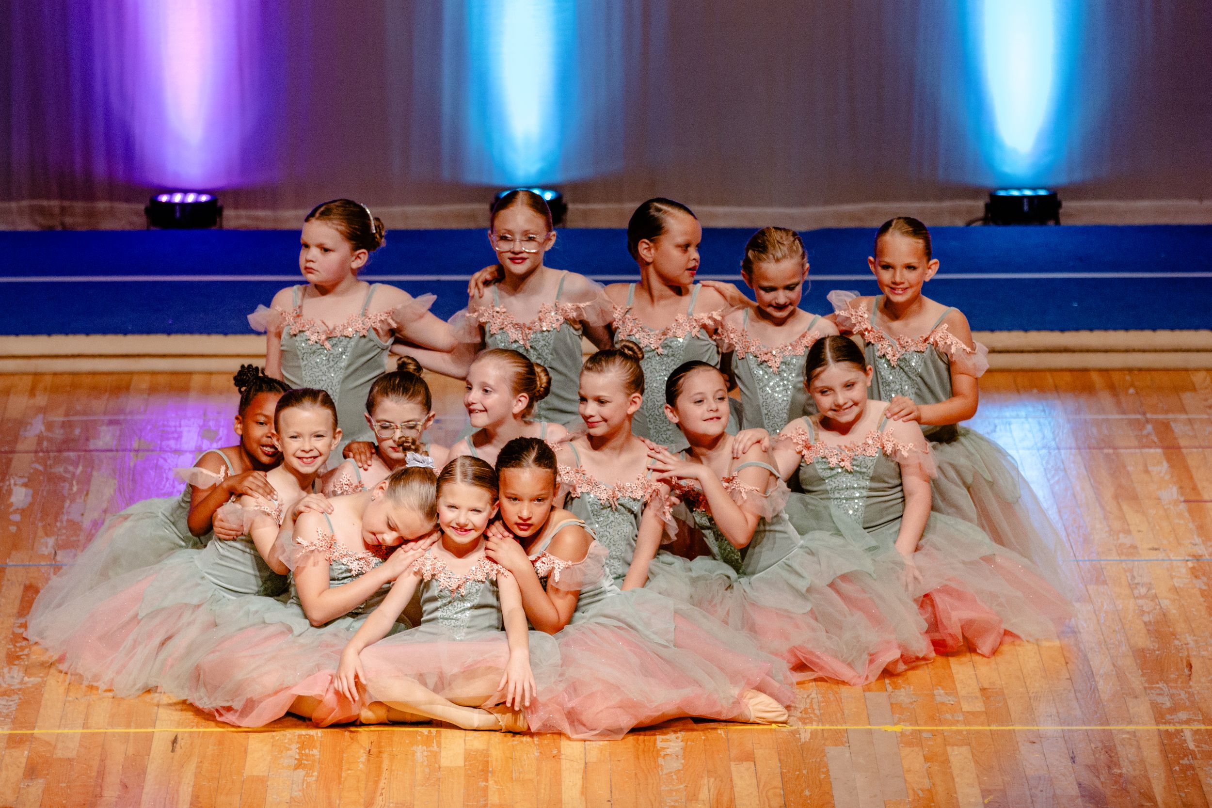 Group of dancers in gray tutus pose together on stage with colorful lighting.