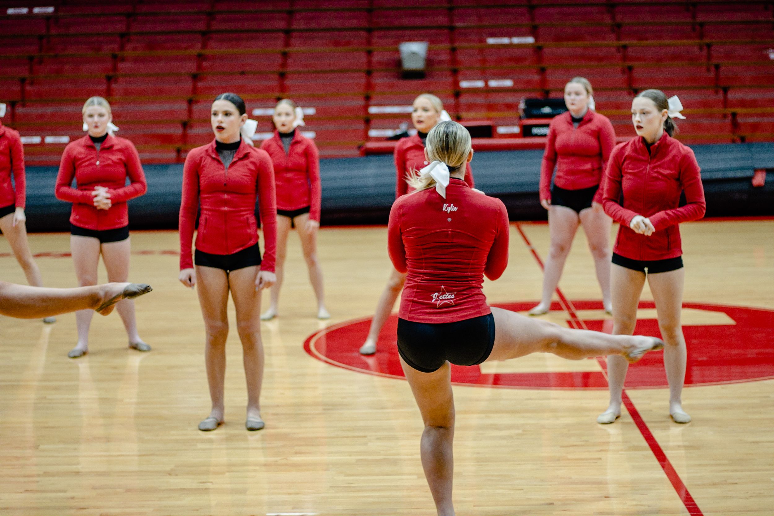 Dance team rehearsing formation movements in red attire on gymnasium floor.