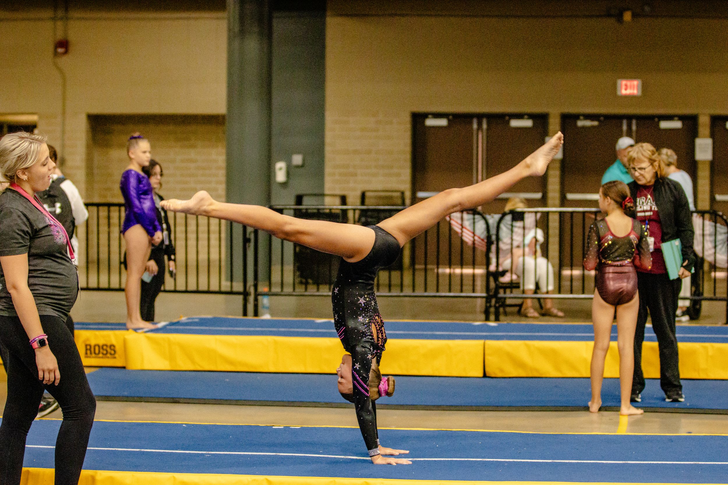 Gymnast performs a split handstand during practice with spotters nearby.
