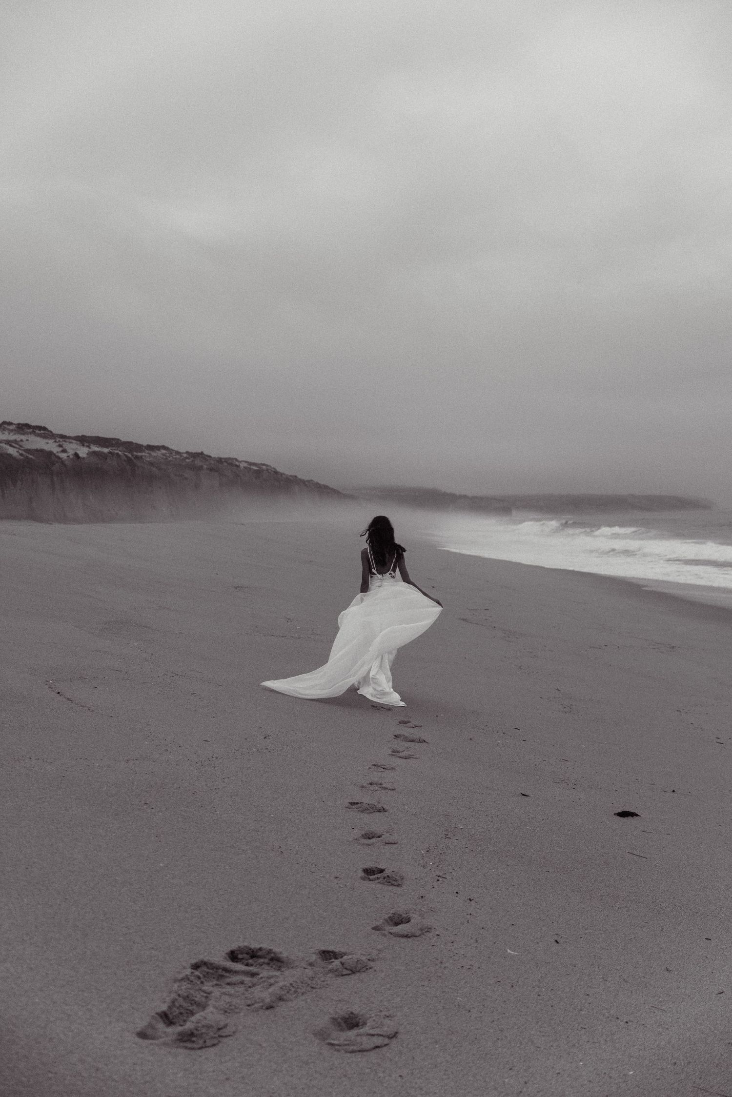 Figure in flowing white wedding dress walking along misty beach leaving footprints in sand in beautiful wedding photo.