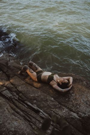 A person relaxes on dark rocks near choppy water on a moody day.