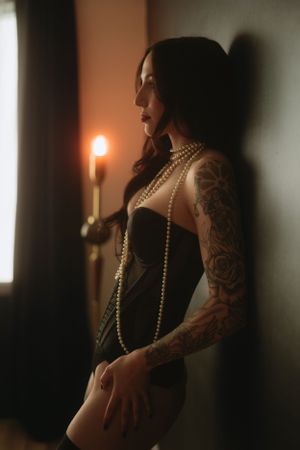 Dark and moody boudoir photo of figure in black corset with pearl necklace and tattoos holding candlelight.