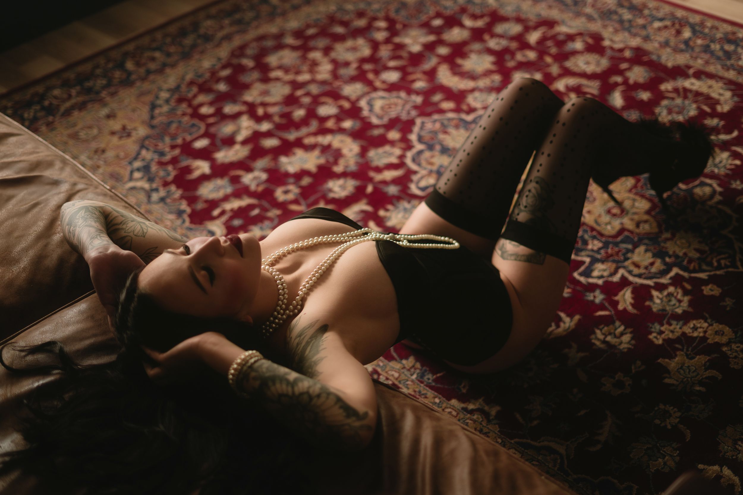 A person lying on an ornate red patterned rug in dark intimate boudoir lighting.