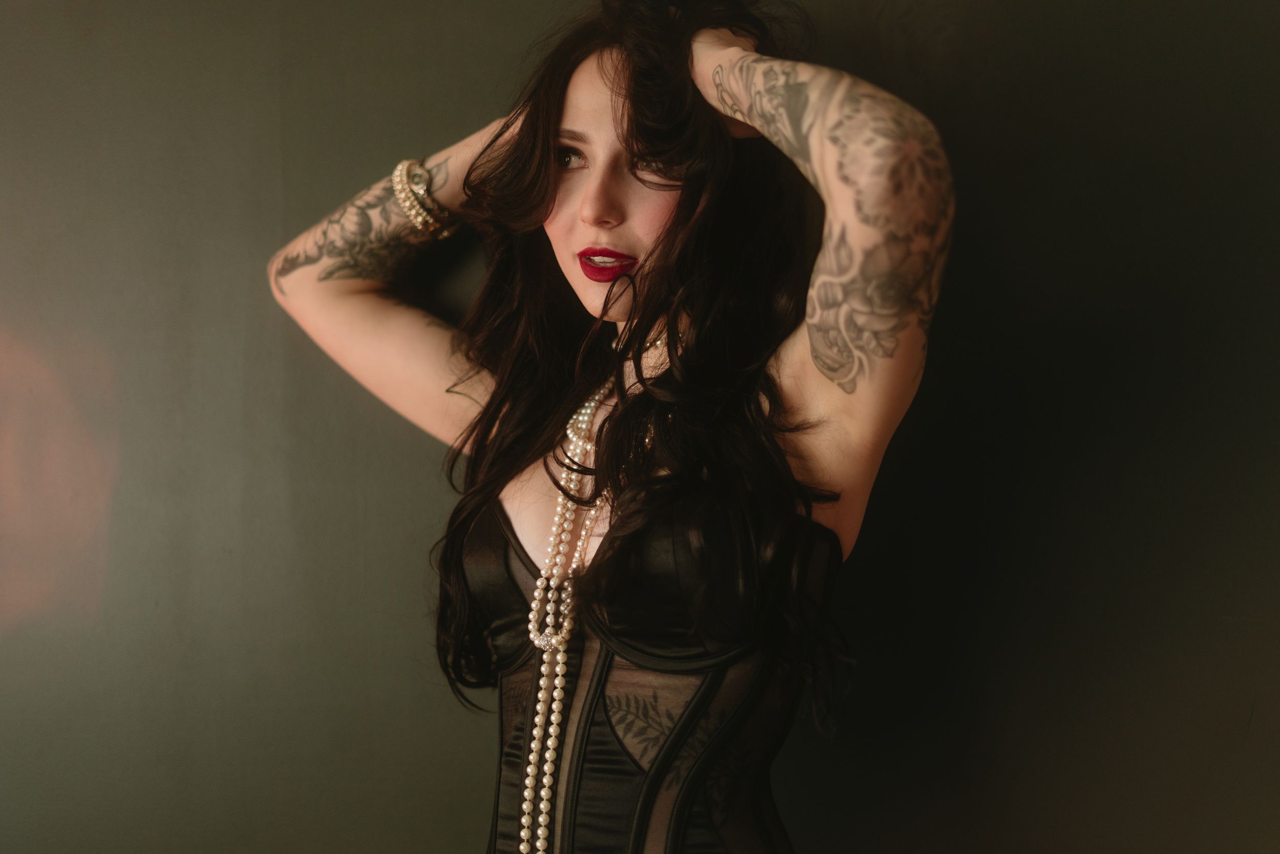 Moody portrait of someone in a black corset with tattoos and long dark hair posing against a dark background.
