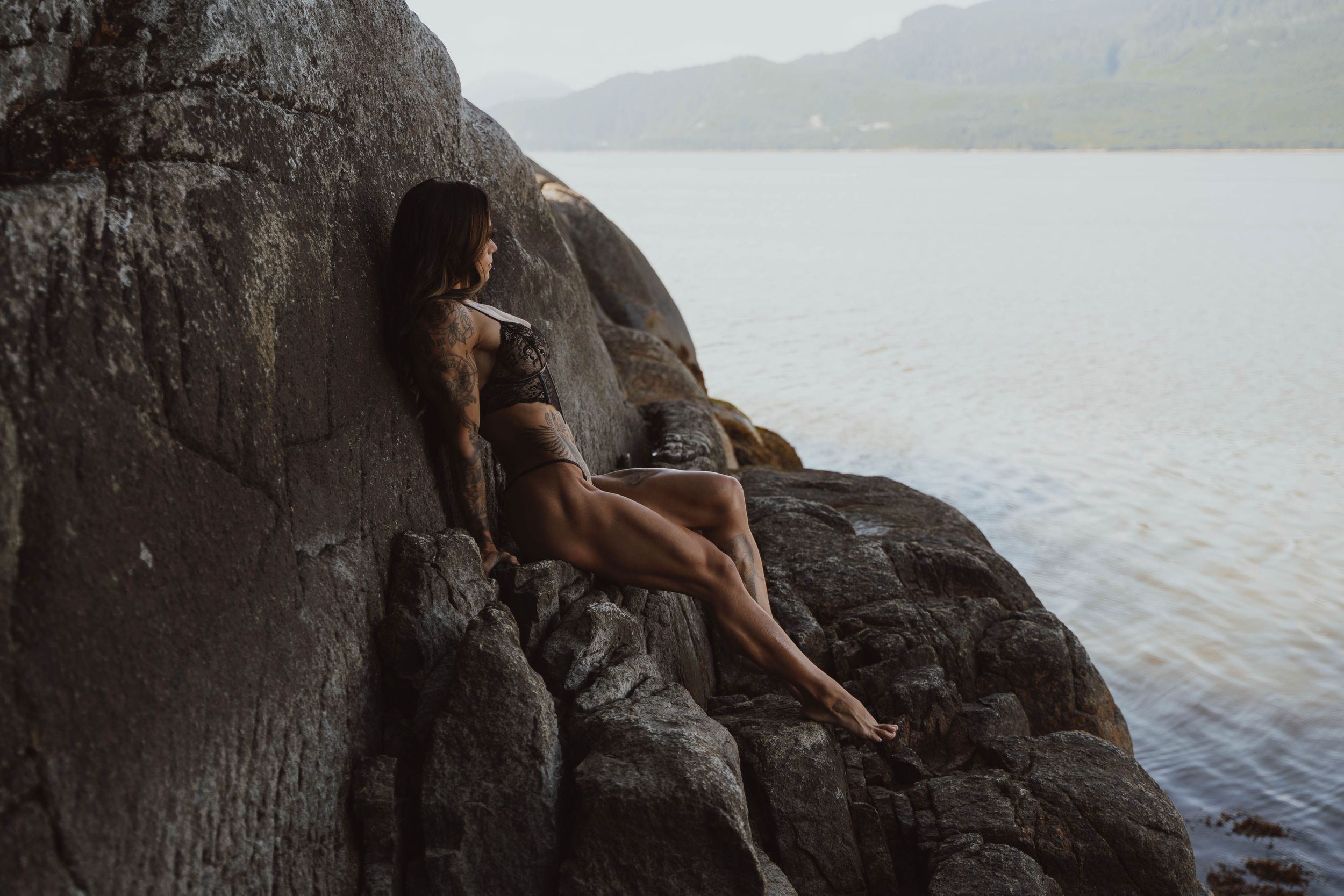 A silhouette rests on coastal rocks overlooking a serene lake and mountains.