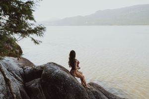 Figure sits contemplatively on dark rocks overlooking a misty lake landscape.