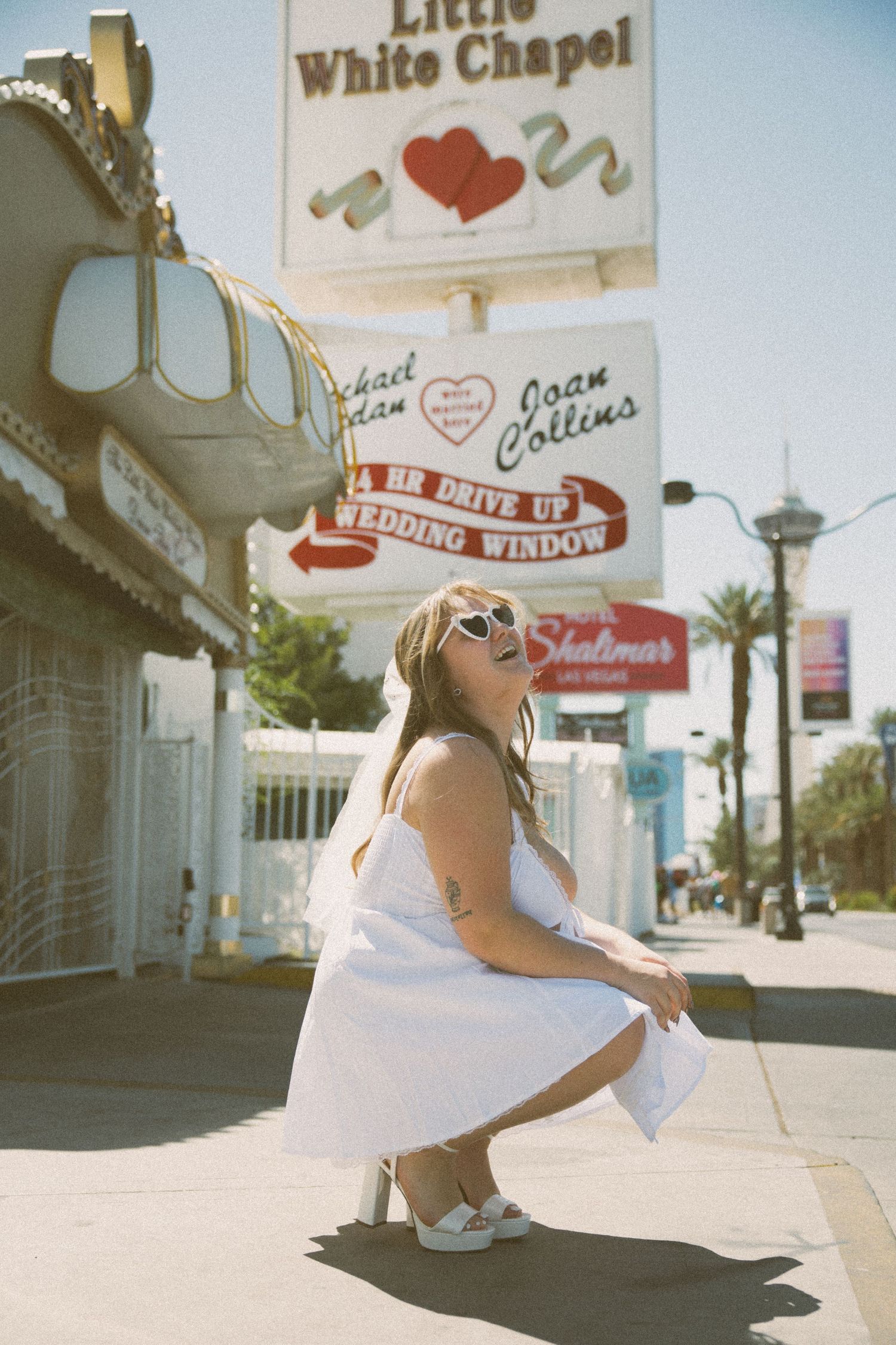 A bride in sunglasses crouches playfully outside the famous Little White Chapel in Las Vegas.
