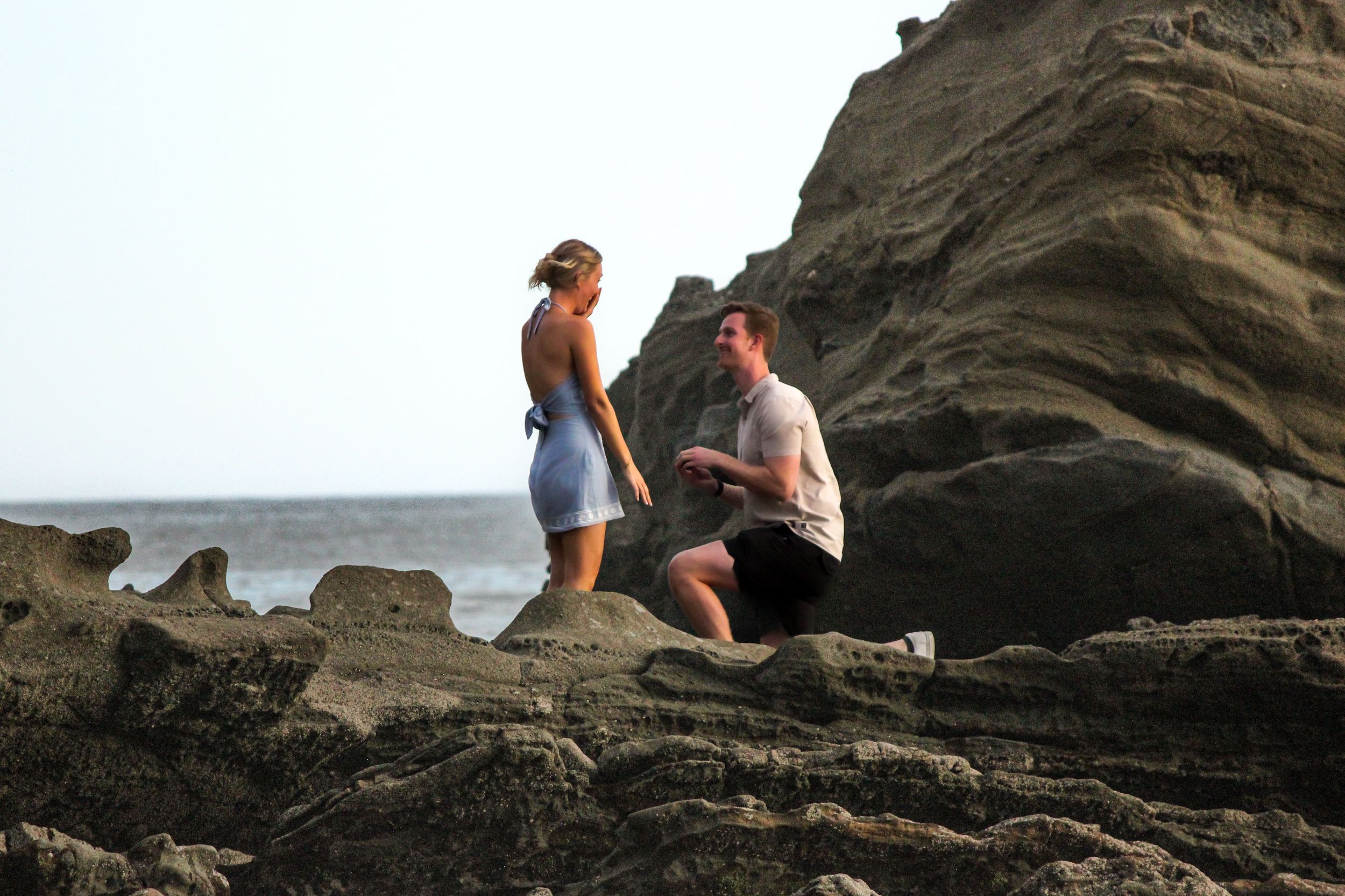 A beach engagement photoshoot taking place on rocks. Man on a knee proposing to a female in a blue dress