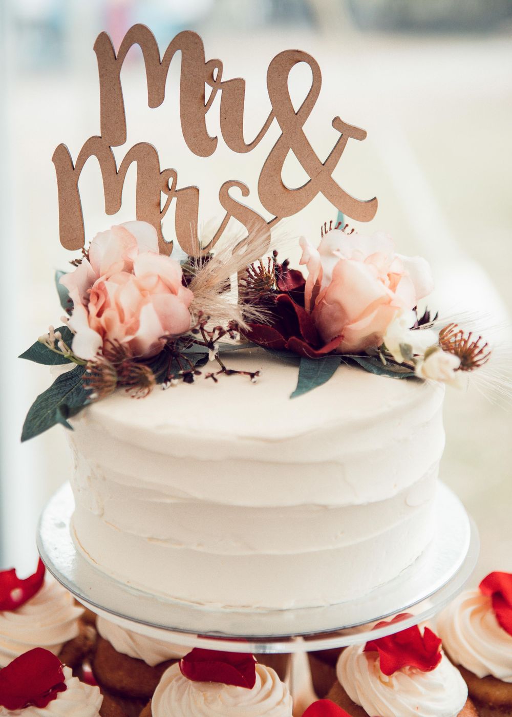 Close up photo of wedding cake topper and flowers surrounded by small cupcakes.