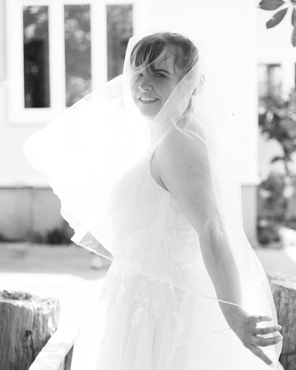 Black and white photo of a smiling bride in a white gown and veil, posing outdoors near a white building.