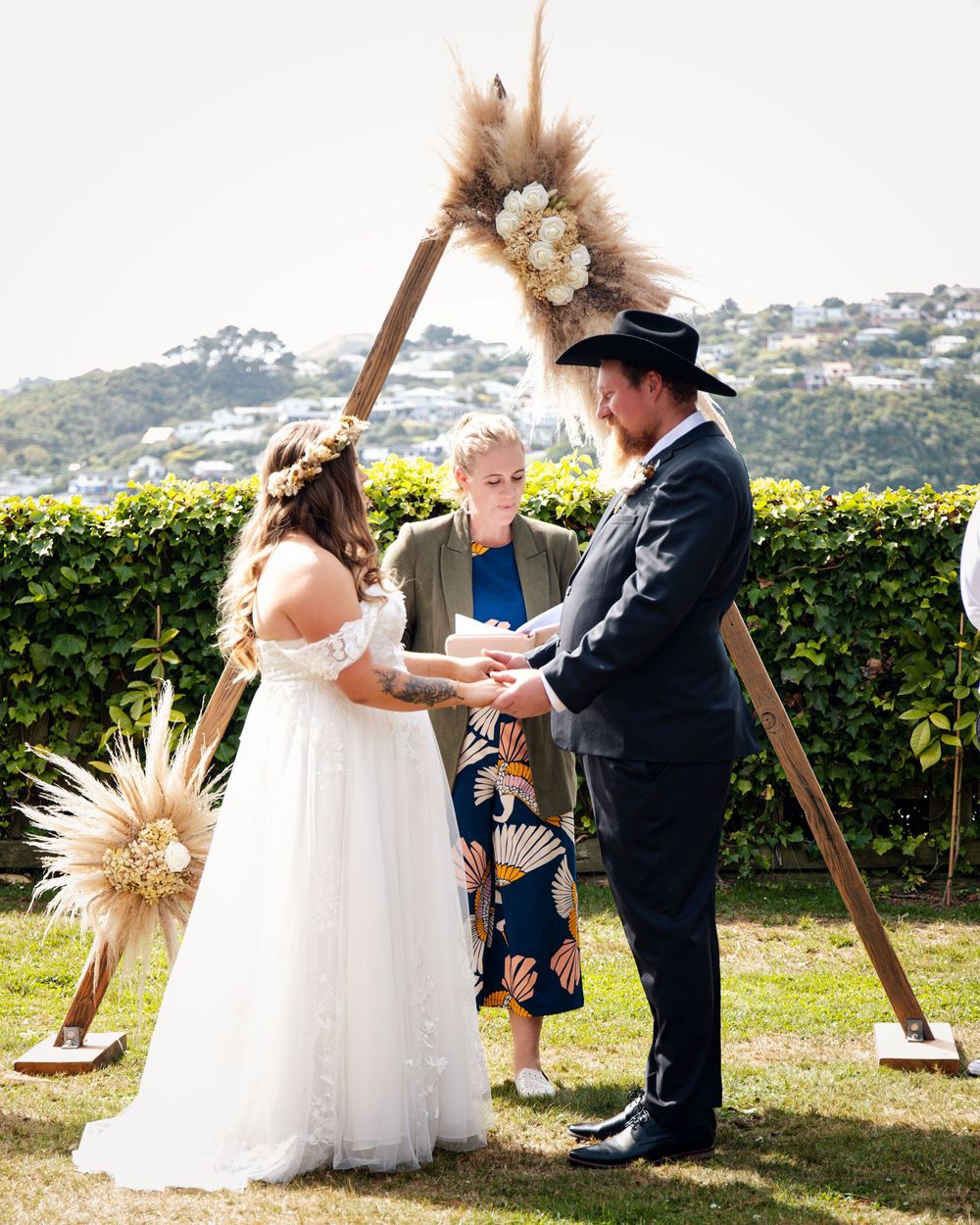 Couple exchanging vows at outdoor boho wedding ceremony with pampas grass triangle arch and green hedge backdrop.