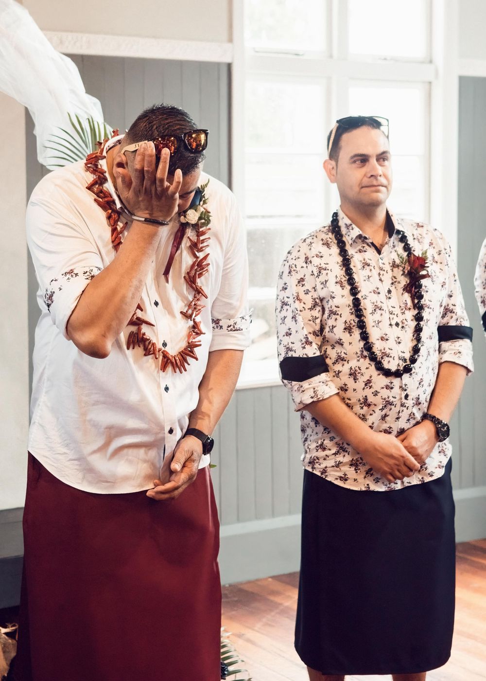 Wedding photo of groom and groomsman dressed in Pasifika clothing, with groom rubbing his eyes as the bride arrives