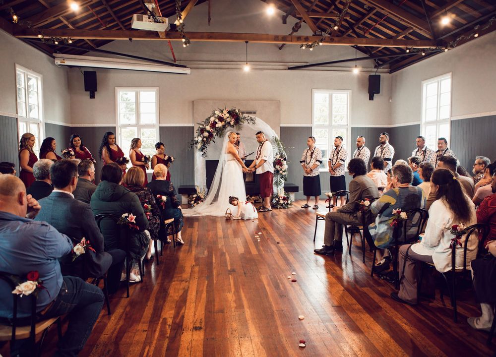 Wide shot of wedding being held indoors, flower girl picking up petals in front of bride and groom.
