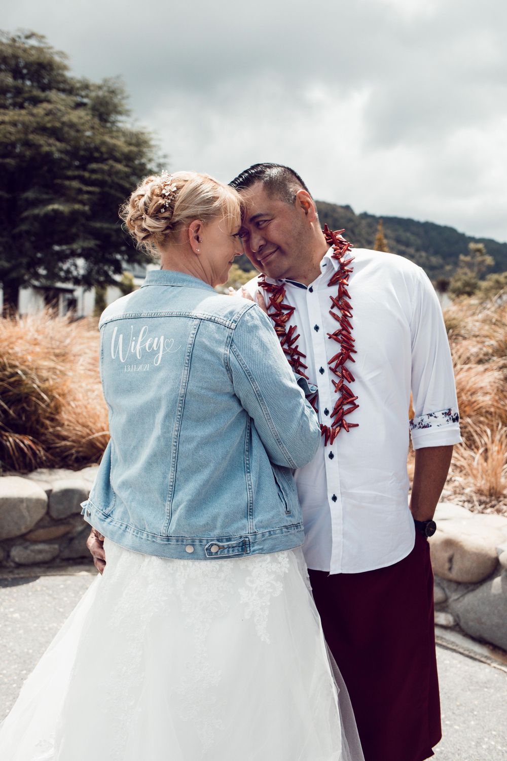 Wedding portrait of bride and groom outside. Bride's back is to the camera and she is wearing a denim jacket saying wife
