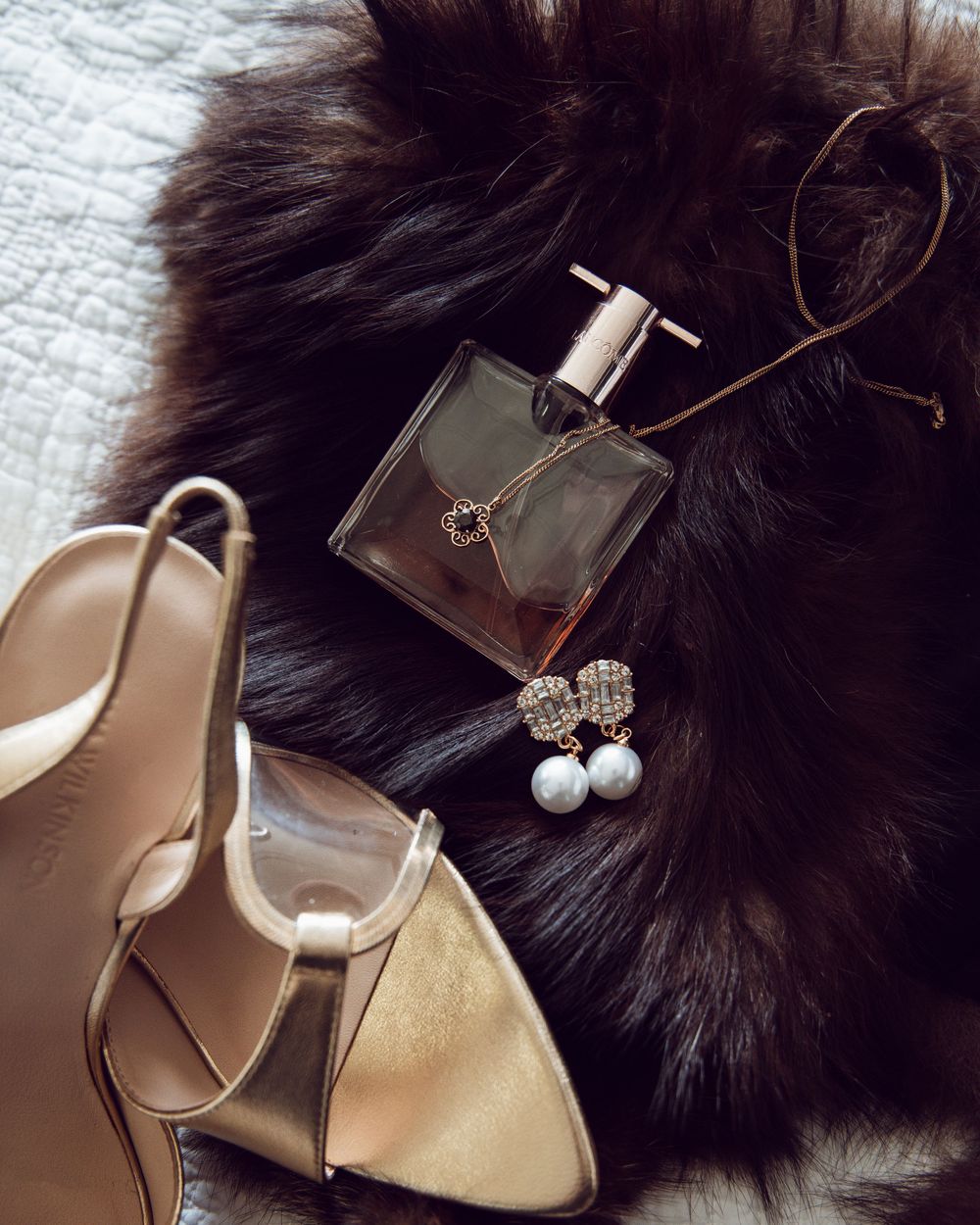 Luxury flat lay with gold heels, perfume bottle, pearl earrings, and fur on white background.