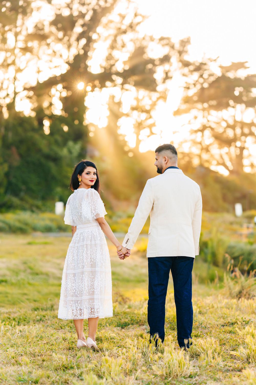 Couple in formal attire holding hands while standing in sunlit meadow at sunset.