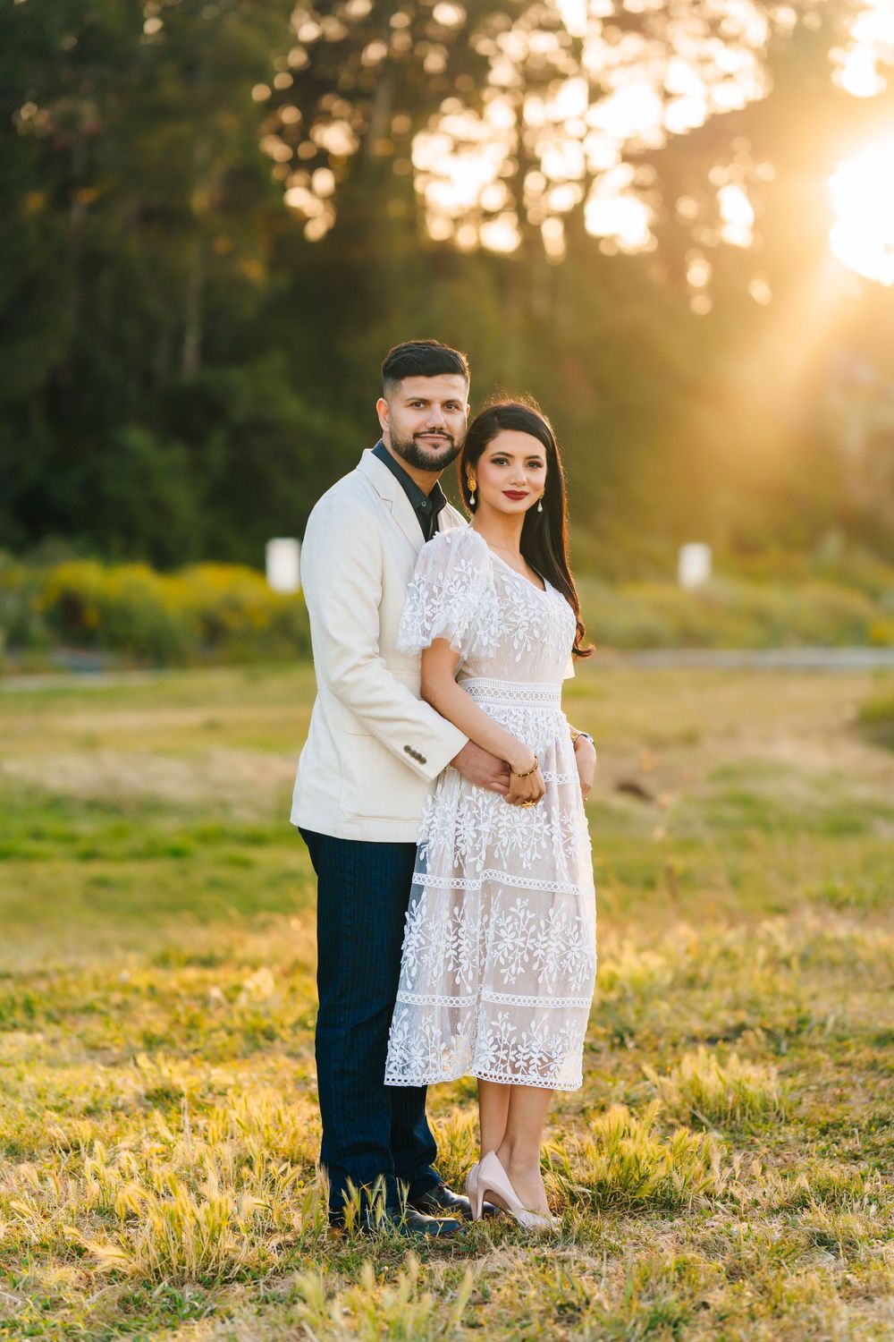 Couple in formal attire pose for portrait in sunlit field during golden hour.