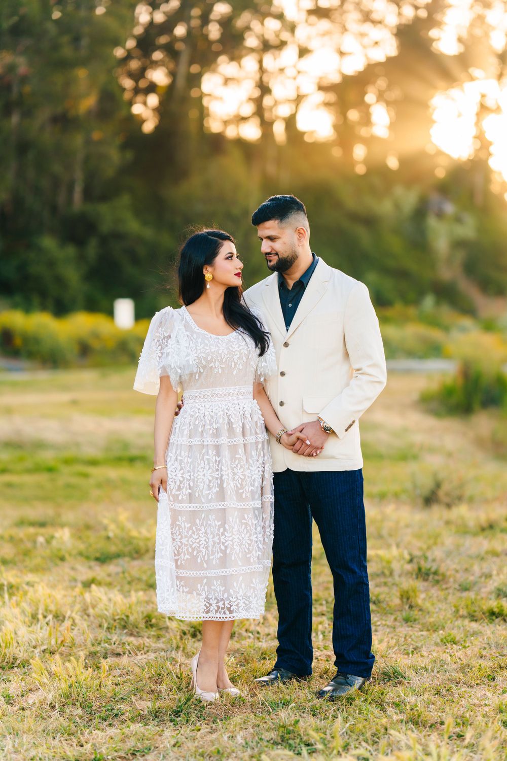 A romantic couple poses in formal attire against a golden sunset in a grassy field.